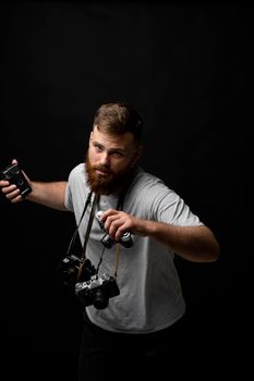 Handsome bearded photographer in a grey t-shirt with a bunch of different cameras in a hands and on a shoulder looking on a camera