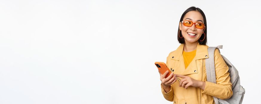 Stylish young asian woman tourist, traveller with backpack and smartphone smiling at camera, posing against white background. Copy space