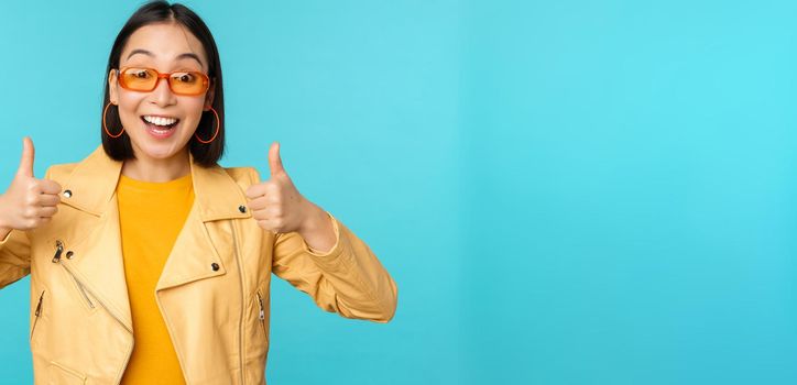 Enthusiastic korean girl looks excited and shows thumbs up, approves smth awesome, excellent choice and quality, stands over blue background.