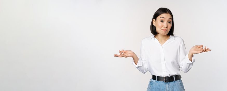 Image of confused asian office manager, office lady shrugging shoulders and looking clueless, standing puzzled against white background.