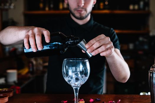 Young and modern bartender, with long dark hair, dressed in black polo shirt, using a stainless steel liquid meter, for the preparation of a mixed drink, on the wooden bar counter... Waiter preparing a mixed drink. Cocktail glass with ice cubes. Gin Tonic. Bar full of cocktail ingredients. Dark background and dramatic lighting.