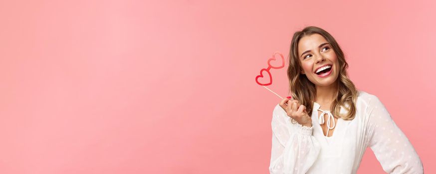 Spring, happiness and celebration concept. Close-up portrait of beautiful lovely blond girl, holding heart-shaped glasses mask and daydreaming, looking up thoughtful with pleased dreamy smile.