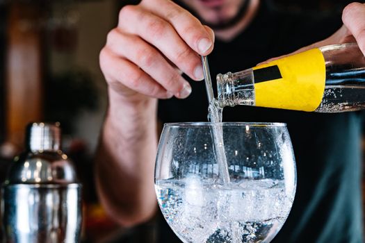 Close-up of a crystal glass with ice, pouring a yellow label soft drink inside for a mixer, on the wooden bar counter of the bar. Waiter preparing a cocktail. Cocktail glass with ice cubes. Gin Tonic. Cocktail shaker. Dark background and dramatic lighting.