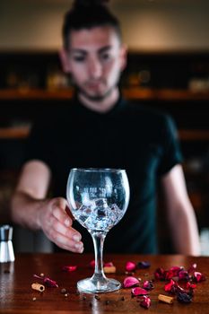 Young and modern waiter, with long dark hair, dressed in black polo shirt, putting a crystal glass with ice cubes on the wooden bar. Waiter preparing cocktail. Cocktail glass with ice cubes. Gin Tonic. Bar full of cocktail ingredients. Dark background and dramatic lighting.