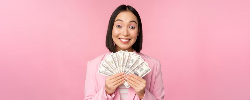 Finance, microcredit and people concept. Happy smiling asian businesswoman showing dollars money, standing in suit against pink background.