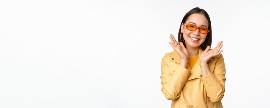 Beautiful asian girl in stylish sunglasses, smiling happy, looking bright and carefree, standing over white background. Copy space