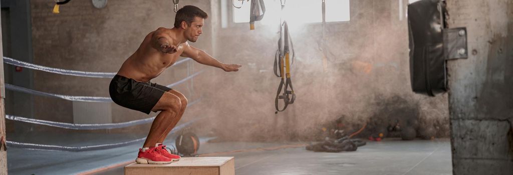 Sporty physically fit man doing step-up exercise on wooden box while doing gym training in sports club