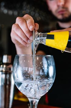 detail of a soft drink with yellow label being poured into a crystal glass for a mixer, on the wooden bar counter of the bar. Waiter preparing a cocktail. Cocktail glass with ice cubes. Gin Tonic. Cocktail shaker. Dark background and dramatic lighting.