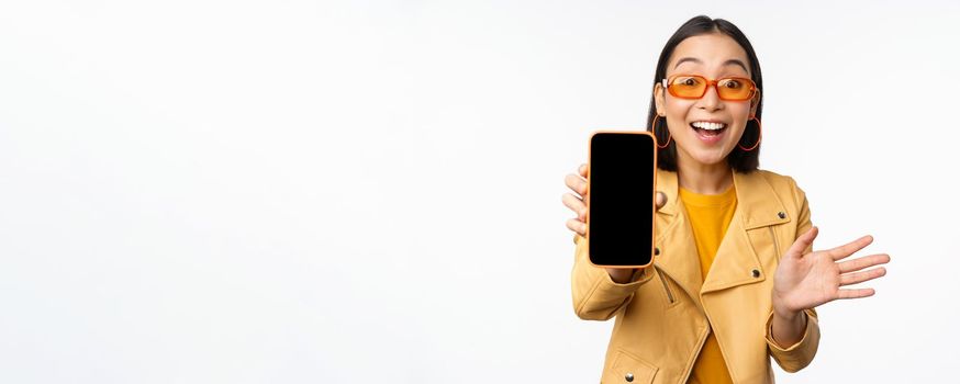 Enthusiastic asian female model, showing smartphone app interface, online store or website on mobile phone screen, standing over white background.