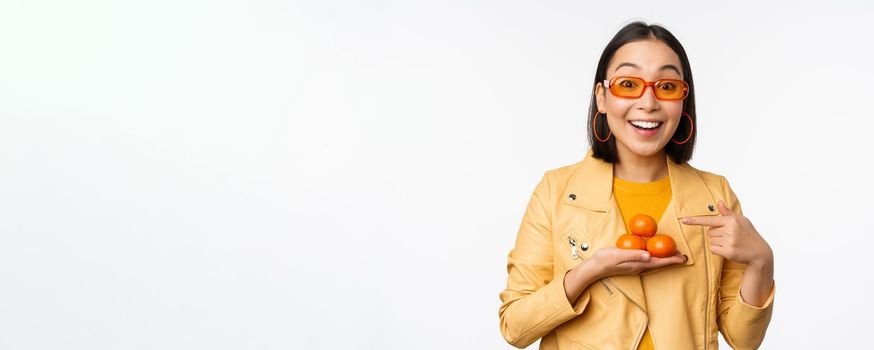 Stylish happy asian girl in sunglasses holding tangerines and smiling, posing against white background. Copy space