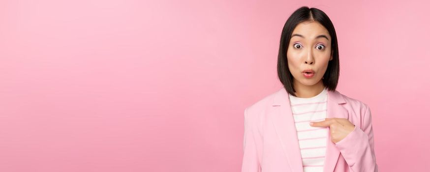 Portrait of asian businesswoman reacts surprised, points at herself with disbelief on face, posing in suit against pink background.