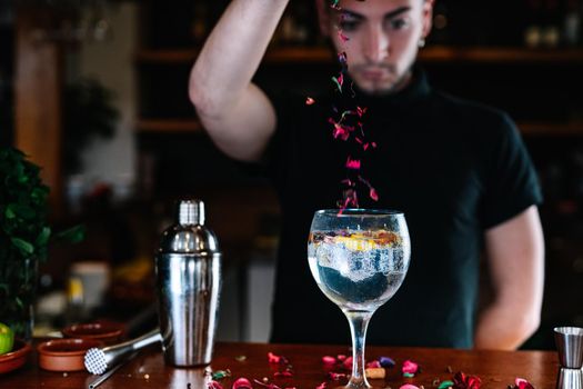 Young, modern waiter, with long dark hair, dressed in black polo shirt, adding toppings, in a crystal glass with ice for a mixer. Waiter preparing a cocktail. Cocktail glass with ice cubes. Gin and tonic. Bar full of cocktail ingredients. Dark background and dramatic lighting.