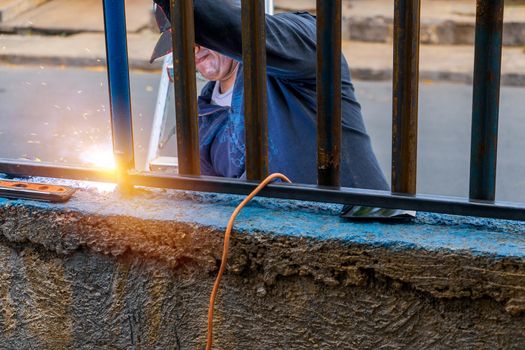 Blue collar worker welding a metal fence unsafely outdoors without protective gear.
