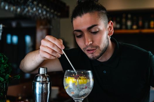 Young and modern bartender with long dark hair, dressed in black polo shirt, stirring a cocktail mix in a crystal glass with a slice of citrus fruit and ice for a cocktail. Waiter preparing a cocktail. Cocktail glass with ice cubes. Gin and tonic. Bar full of cocktail ingredients. Dark background and dramatic lighting.