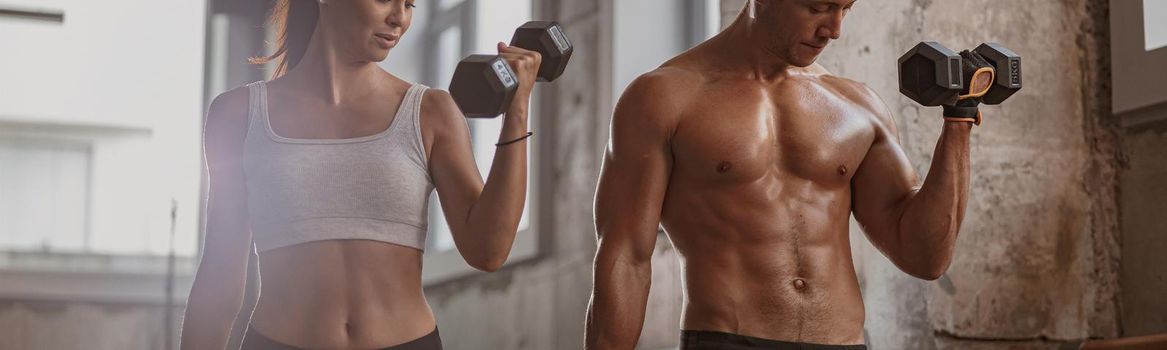 Slender young female and athletic man holding black dumbbells and doing hand exercise together in the gym