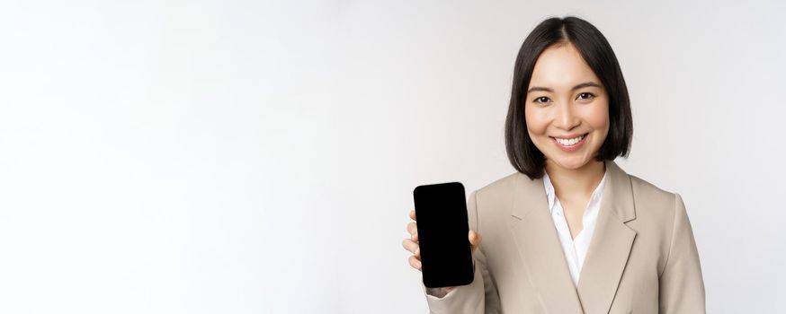 Portrait of corporate asian woman showing smartphone app interface, mobile phone screen, standing over white background.