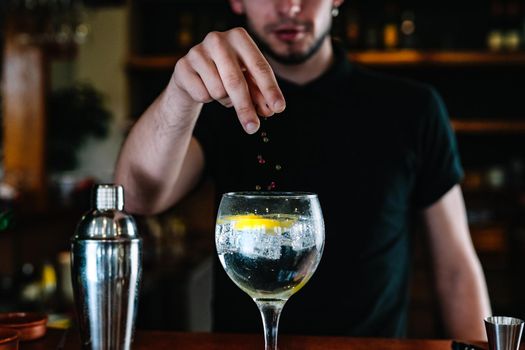 Young and modern waiter, with long dark hair, dressed in black polo shirt, adding pepper garnishes, in a crystal glass with ice for a mixer. Waiter preparing a cocktail. Cocktail glass with ice cubes. Gin Tonic. Bar full of cocktail ingredients. Dark background and dramatic lighting.