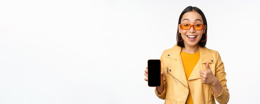 Beautiful korean girl, asian woman in sunglasses, showing smartphone app interface, thumbs up, recommending mobile phone application, white background.