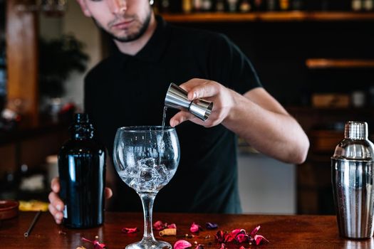 Young and modern bartender, with long dark hair, dressed in black polo shirt, pouring liquor into a crystal glass to prepare a mixed drink, on the wooden bar counter... Waiter preparing a cocktail. Cocktail glass with ice cubes. Gin Tonic. Bar full of cocktail ingredients. Dark background and dramatic lighting.