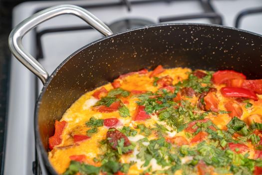 Fried egg with vegetables and herbs in black frying pan on white stove. Side view