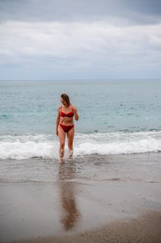 A middle-aged woman with a good figure in a red swimsuit on a pebble beach, running along the shore in the foam of the waves.