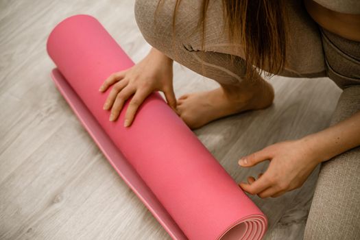 A young woman rolls a pink fitness or yoga mat before or after exercising, exercising at home in the living room or in a yoga studio. Healthy habits, keep fit, weight loss concept. Closeup photo.