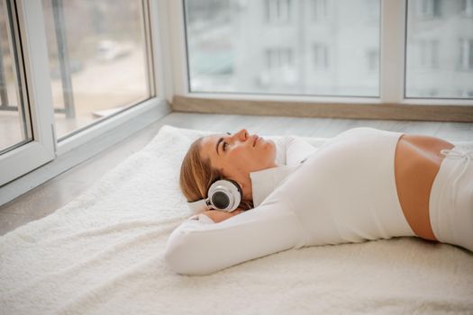 Side view portrait of relaxed woman listening to music with headphones lying on carpet at home. She is dressed in a white tracksuit