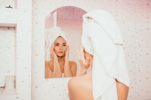 Young beautiful woman using face cream moisturizing lotion after bath. Pretty attractive girl in a towel on her head stands in front of a mirror in a home bathroom. Daily hygiene and skin care.