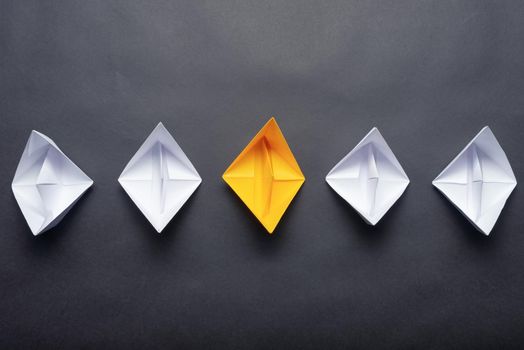 Row of paper ships on black background. Business concept of creative innovation and leadership. Flat lay yellow origami boat in group of white boats. Social marketing layout with copy space.