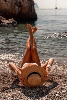 A beautiful middle-aged woman lies on the beach with her feet to the sky, covering her body with a wide-brimmed straw hat. She is sunbathing on the ocean. Vacation, travel, vacation concept