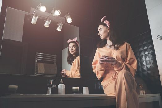 Portrait view of beautiful young woman getting ready in home bathroom decorated with black tiles. moisturising cream. female cosmetics, mirror face reflection.