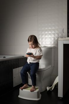 A girl dressed in a blue jeance and white t-shirt playing with a mobile phone in the bathroom.