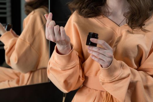 Woman is holding an open jar of cream and applies a small amount of the product to her hand. Concept of skin care, testing and selection of high-quality natural and organic cosmetics. Closeup