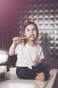 A small child in blue jeanse and white t-shirt brushes his teeth with a bamboo toothbrush. eco friendly. Black tiles on a wall. White sink