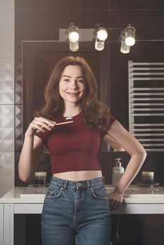 Woman brushing her teeth. Young woman in a burgundy top and jeanse with bamboo brush in front of the mirror. Bathroom with a black tiles