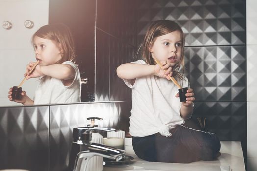 A small child in blue jeanse and white t-shirt brushes his teeth with a bamboo toothbrush. eco friendly. Black tiles on a wall. White sink