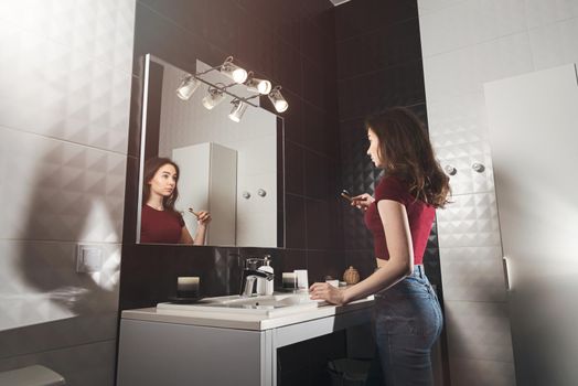 Woman brushing her teeth. Young woman in a burgundy top and jeanse with bamboo brush in front of the mirror. Bathroom with a black tiles
