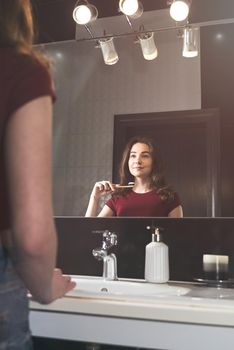 Woman brushing her teeth. Young woman in a burgundy top and jeanse with bamboo brush in front of the mirror. Bathroom with a black tiles