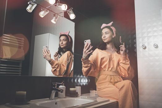 beautiful young woman communicating by the phone and doing her hair in a stylish bathroom. dressed in an orange sports suit and a bow and sits near the mirror