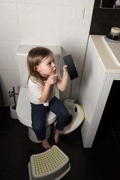 A girl dressed in a blue jeance and white t-shirt playing with a mobile phone in the bathroom.