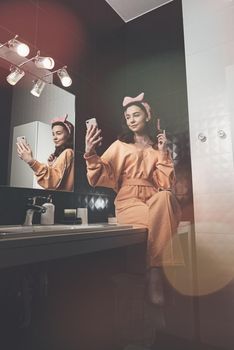 beautiful young woman communicating by the phone and doing her hair in a stylish bathroom. dressed in an orange sports suit and a bow and sits near the mirror