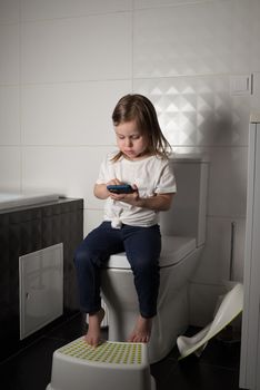 A girl dressed in a blue jeance and white t-shirt playing with a mobile phone in the bathroom.