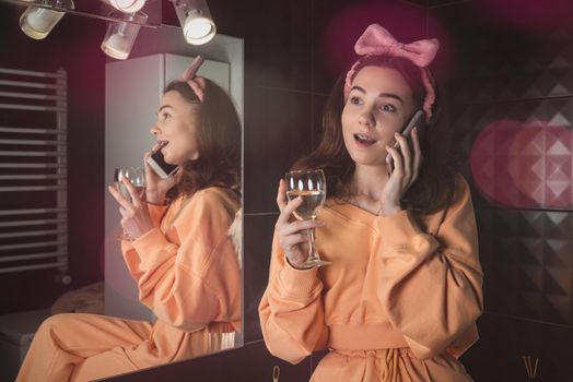 Pretty funny woman with mobile phone in the bathroom whit a glass of water in a hand. Woman wearing orange pajamas and a bow on his head