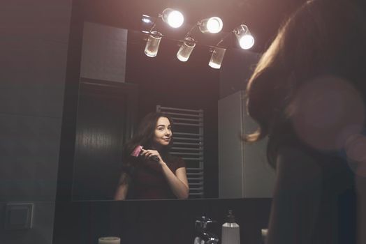 Young woman brushing hair in front of a bathroom mirror. Black tiles on a wall