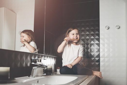 A small child in blue jeanse and white t-shirt brushes his teeth with a bamboo toothbrush. eco friendly. Black tiles on a wall. White sink