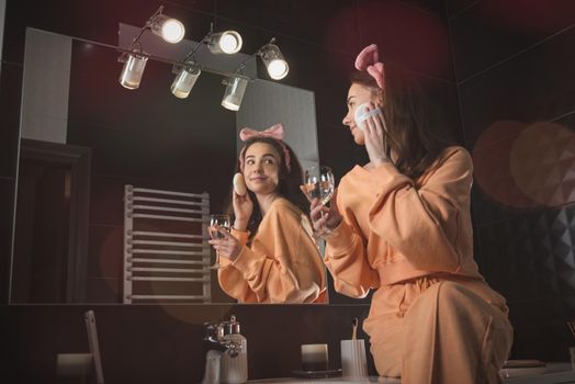 Pretty funny woman with mobile phone in the bathroom whit a glass of water in a hand. Woman wearing orange pajamas and a bow on his head