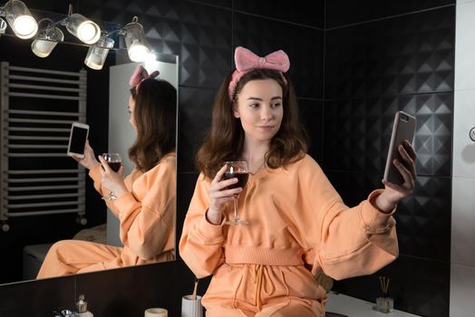 Attractive young woman in a bathroom communicating on a smartphone and drinking wine. Relaxation and leisure concept. girl dressed in orange pajamas