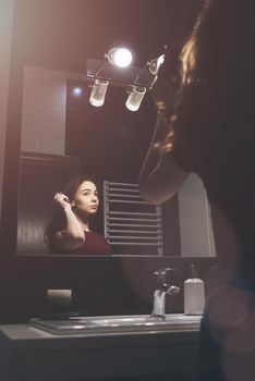 Young woman brushing hair in front of a bathroom mirror. Black tiles on a wall