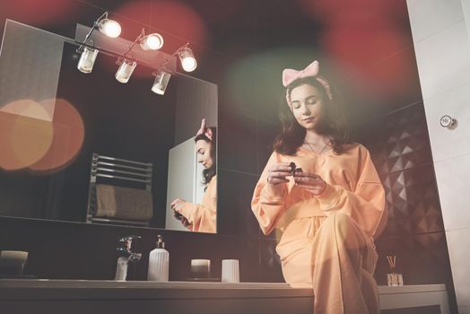 Portrait view of beautiful young woman getting ready in home bathroom decorated with black tiles. moisturising cream. female cosmetics, mirror face reflection.