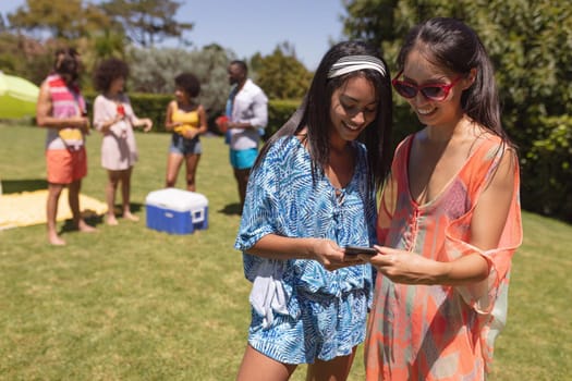 Two mixed race female friends using smartphone at a pool party. Hanging out and relaxing outdoors in summer.